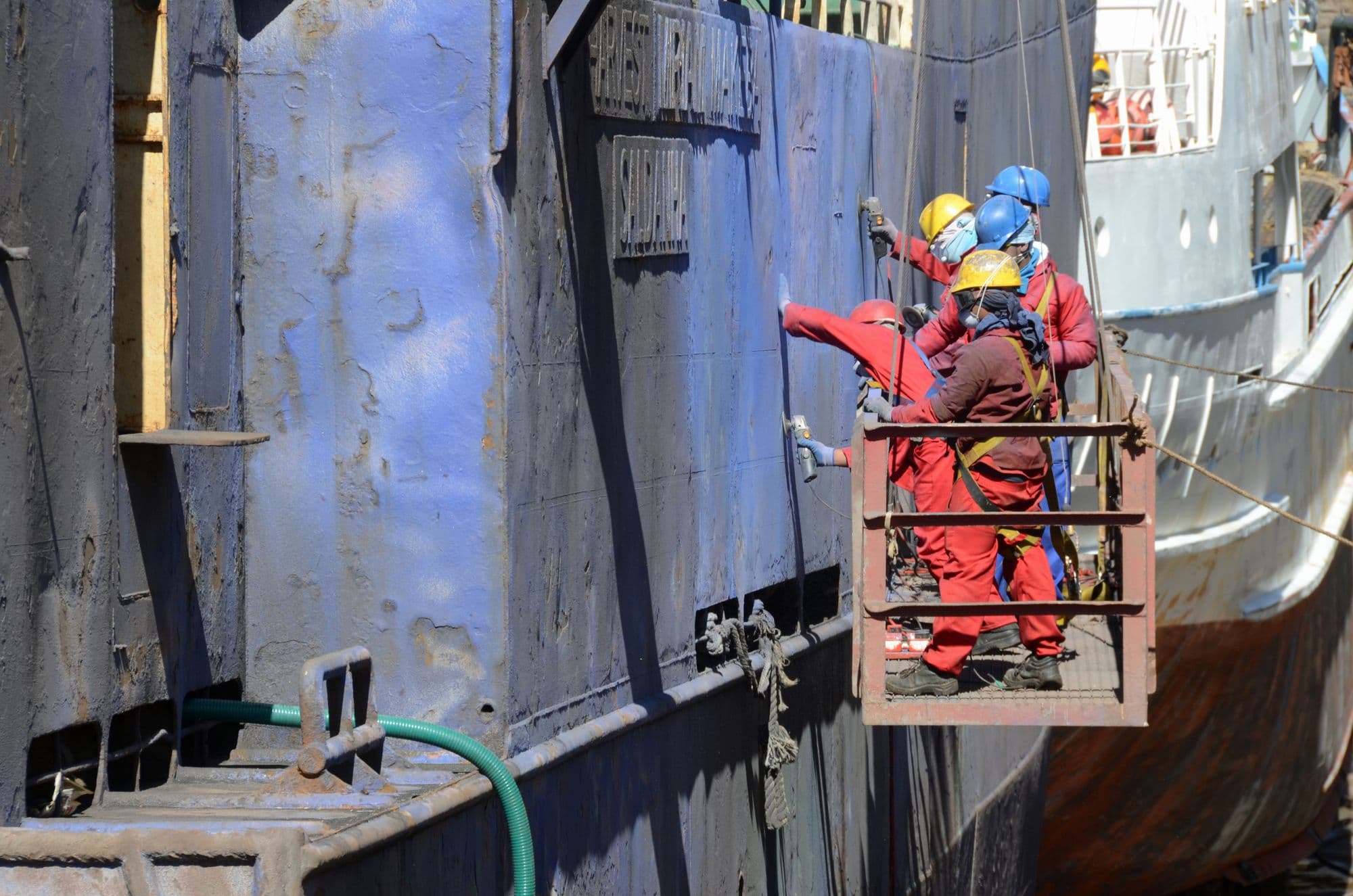 Workers in safety harnesses repairing a ship hull from a suspended platform